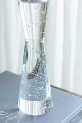 A clear glass hourglass-shaped vase filled with water and small bubbles, resting on a gray surface with a soft white curtain background.