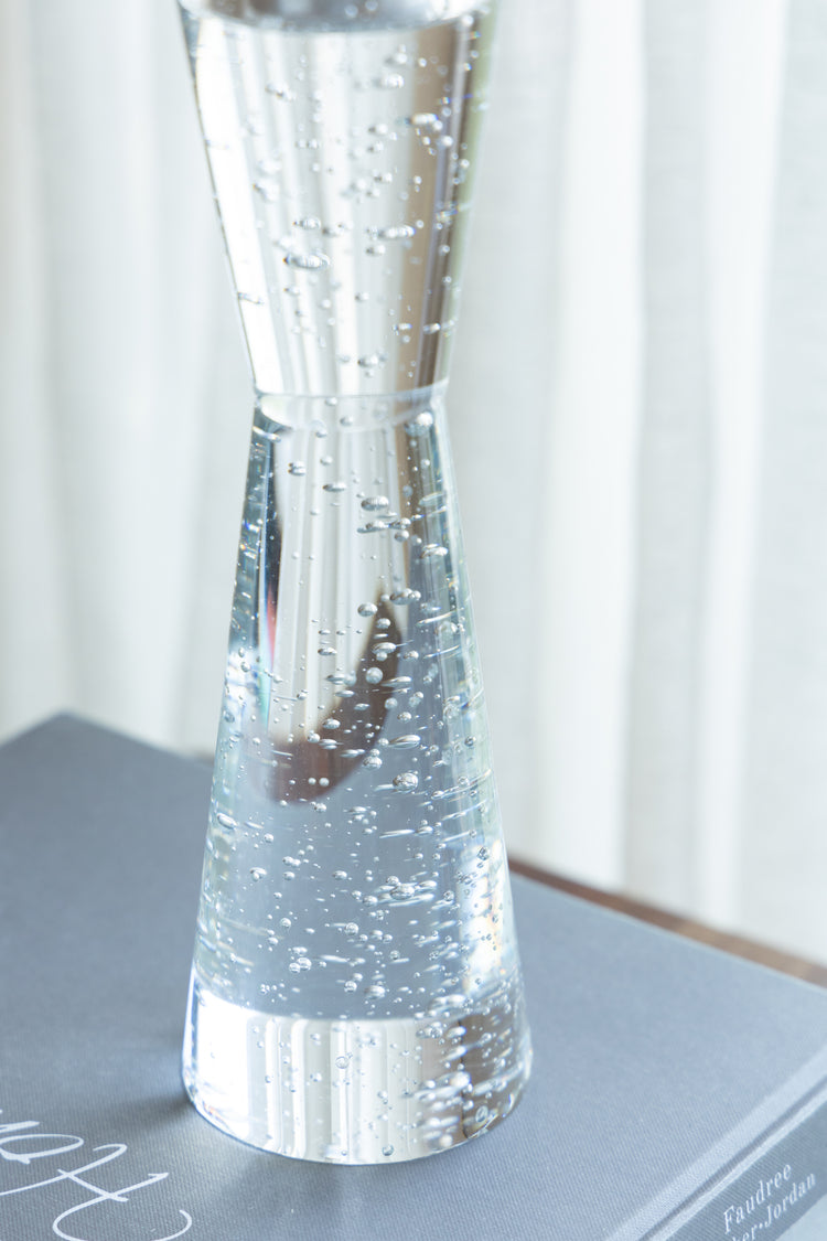 A clear glass hourglass-shaped vase filled with water and small bubbles, resting on a gray surface with a soft white curtain background.