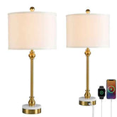 Two matching table lamps with white drum shades, gold-toned stems and finials, and round white marble-look bases, shown on a white background. One lamp is fully visible, and the other is shown with a power strip and charging cables connected, alongside a smartwatch and smartphone screen.