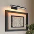 A modern wall-mounted linear light fixture with a black frame and warm white LED illumination, shown above a framed abstract artwork on a beige wall with a green plant in the lower right corner.