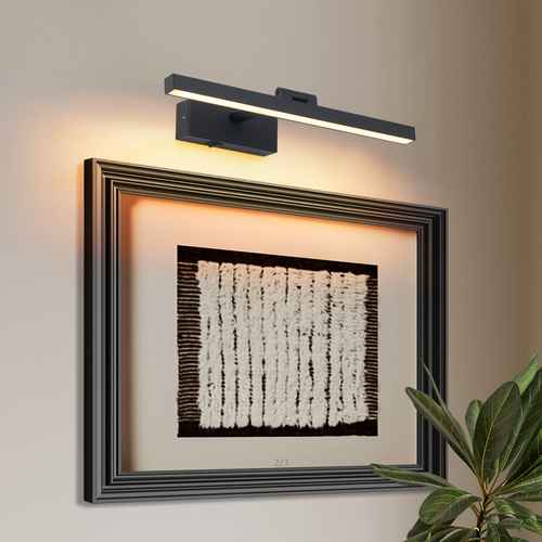 A modern wall-mounted linear light fixture with a black frame and warm white LED illumination, shown above a framed abstract artwork on a beige wall with a green plant in the lower right corner.
