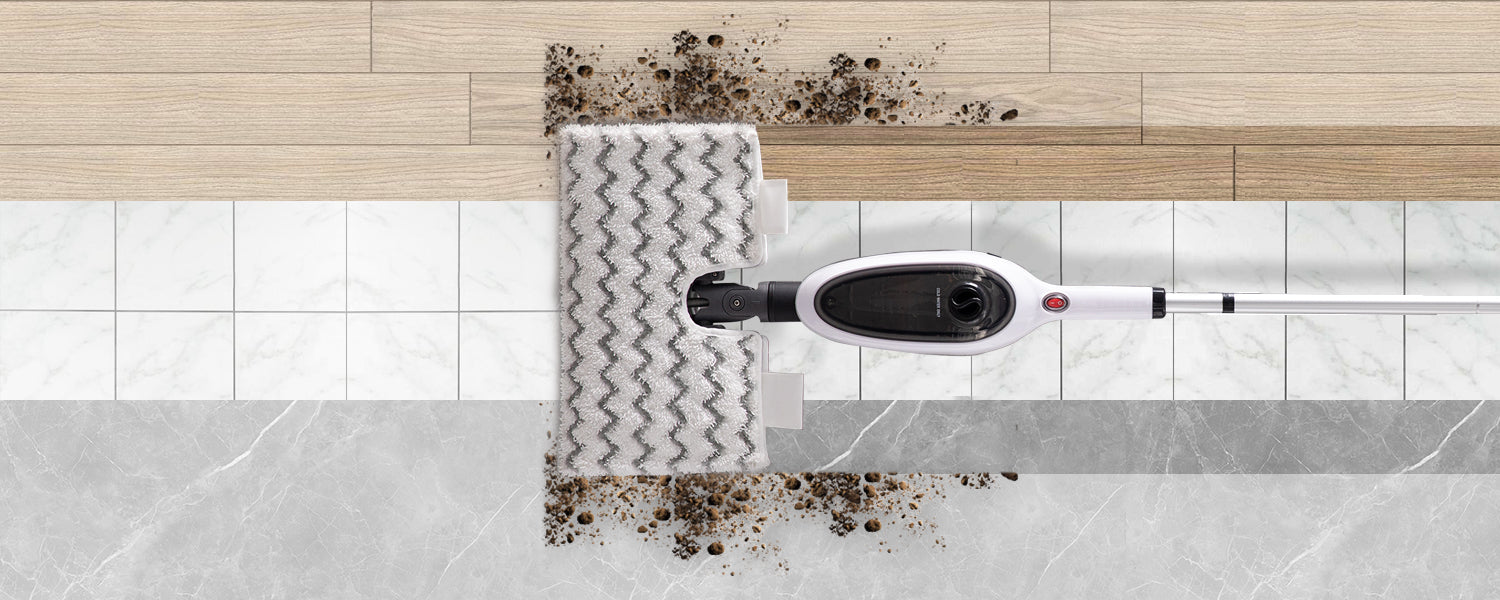 A wet-dry floor cleaner shown from above, with a white and black head lifting dark dirt over a tiled floor transition between wood and marble surfaces.