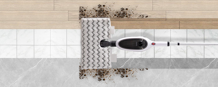 A wet-dry floor cleaner shown from above, with a white and black head lifting dark dirt over a tiled floor transition between wood and marble surfaces.