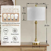 A table lamp with a white drum shade, gold-toned stem and base details, and a white round base, shown on a wooden table with overlaid dimension labels (13 in, 9.5 in, 28.