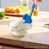 A blue and green plastic scoop or funnel-shaped measuring tool being used to transfer white powder into a clear glass bowl on a wooden countertop, with lemons and jars in the background.