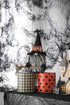 A Halloween-themed gnome decoration with a tall black hat, orange plaid pattern, and long gray beard, posed on top of two cylindrical containers against a smoky black-and-white backdrop.