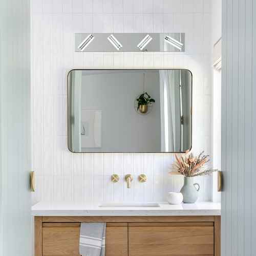A modern rectangular bathroom mirror with a thin dark frame, mounted on a white beadboard wall above a white countertop with gold faucet hardware. A horizontal light fixture with four rectangular bulbs is mounted above the mirror.