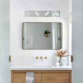 A modern rectangular bathroom mirror with a thin dark frame, mounted on a white beadboard wall above a white countertop with gold faucet hardware. A horizontal light fixture with four rectangular bulbs is mounted above the mirror.