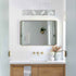 A modern rectangular bathroom mirror with a thin dark frame, mounted on a white beadboard wall above a white countertop with gold faucet hardware. A horizontal light fixture with four rectangular bulbs is mounted above the mirror.
