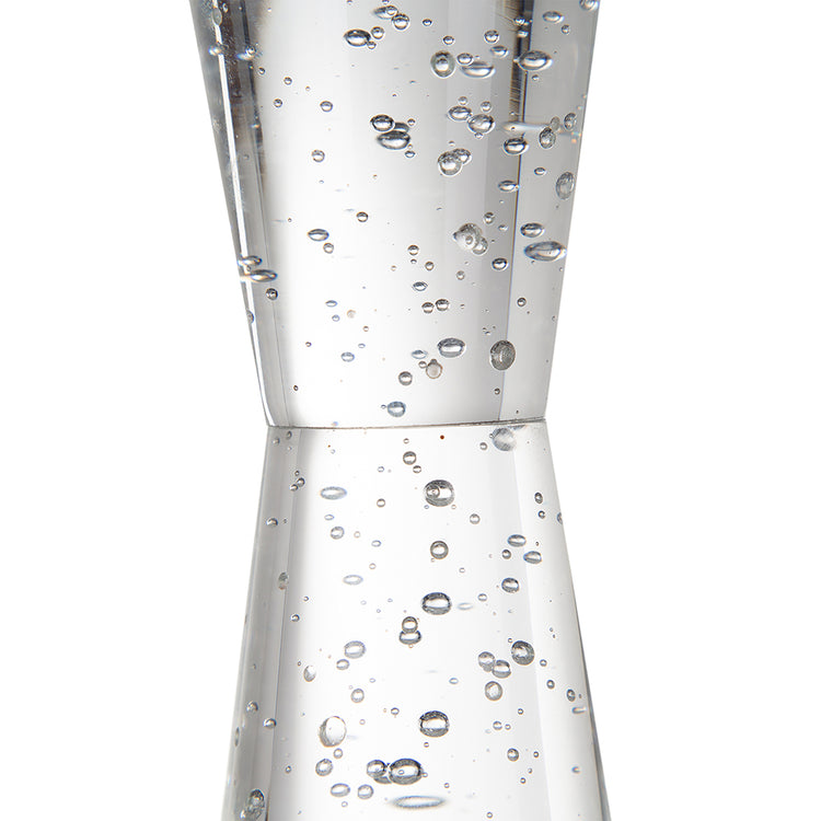 A clear glass jigger with a double-cone shape, filled with small bubbles and photographed against a plain white background.