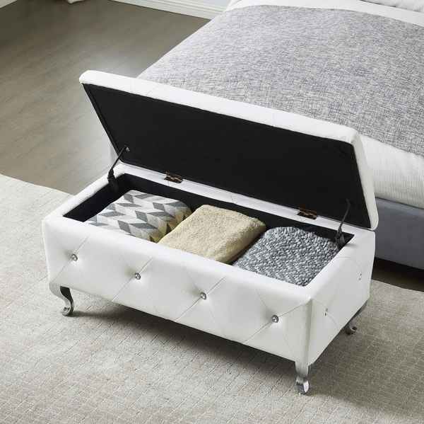A white tufted storage bench with silver legs, shown open on a light rug in front of a bed. The bench is styled with three folded throw pillows in gray, beige, and black-and-white patterns.