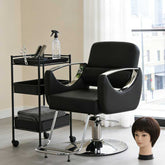 Black salon chair with a cart and mannequin head in a room with large windows.