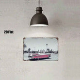 A rectangular wall art print with a distressed vintage border hangs on a textured light gray wall beneath a dark dome pendant light. The print shows a black-and-white city street scene with a faded pink convertible car.
