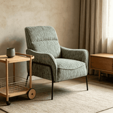 A modern upholstered accent chair in textured sage green fabric with black metal legs, styled in a warm, minimalist room next to a wooden side table.