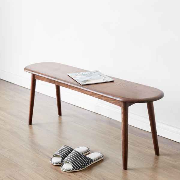A long, low wooden bench with tapered legs sits on a light wood floor against a white wall. A magazine rests on the bench, and a pair of striped slip-on slippers is placed in front.