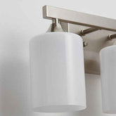 A close-up of a modern wall-mounted light fixture with two frosted white cylindrical shades and a brushed metal bar in a warm silver finish.