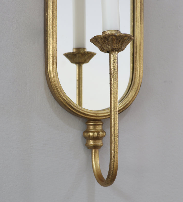 A close-up of a gold wall sconce with an oval mirror and candle-style light fixture, mounted on a light gray wall.