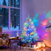 A festive tinsel Christmas tree decorated with multicolored lights, styled in a cozy living room with wrapped gifts, candles, and a window showing a snowy outdoor view.